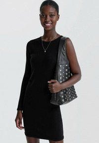 Jeune femme portant une robe noire à une manche, tenant un sac à main en cuir noir clouté, souriante sur un fond uni.