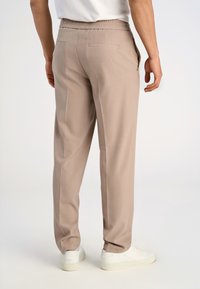 Mężczyzna w beżowych spodniach prostych z elastycznym pasem i białych trampkach, stojący na tle jasnej, jednolitej ściany.