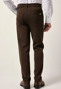 Hombre con pantalones marrones con bolsillos traseros, camisa blanca y zapatos de vestir marrones, de pie contra un fondo liso.