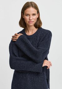 Pull oversized bleu marine en tricot texturé, avec un col côtelé et de longues manches, vu avec les bras croisés sur la poitrine.