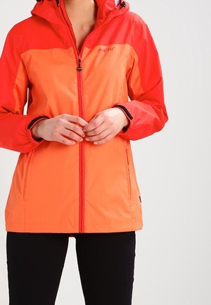 Donna che indossa una giacca antipioggia bicolore rossa e arancione con cappuccio, che chiude la cerniera frontale, con pantaloni neri visibili.