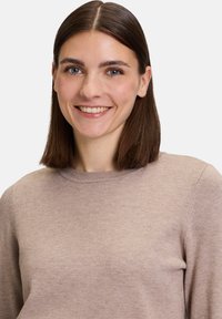 Maglione lavorato a maglia color marrone chiaro, con scollatura tonda, maniche lunghe e una texture morbida, indossato da un modello su uno sfondo bianco.