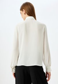 Blusa blanca de manga larga que presenta una textura suave y sedosa con un collar clásico y puños fruncidos, combinada con pantalones negros.