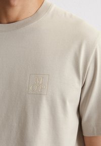 Beige Baumwoll-T-Shirt mit einem dezenten gestickten Logo in einem Quadrat. Kurze Ärmel und klassischer Rundhalsausschnitt. Weicher Stoff.