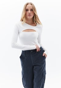Weißes Langarmoberteil mit einem Cut-Out-Detail an der Vorderseite, kombiniert mit dunkelblauen Cargo-Hosen mit Seitentaschen und einer lockeren Passform.