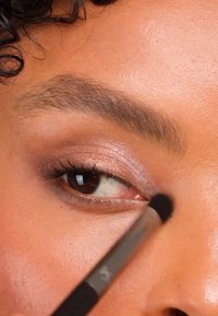 Close-up van een oog met zachte, glinsterende oogschaduw in rozetinten, gedefinieerde wimpers en een make-upkwast die product aanbrengt langs het ooglid.