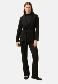 Černé sako s opaskem, velkými klopami a předními kapsami, doplněné o úzké černé kalhoty. Modelka drží malou černou a zlatou kabelku.