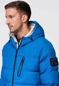 Sininen toppatakki hupulla, jossa vetoketjullinen rintatasku, mustat vetoketjut ja hihassa sininen korostuslaatta. Sileä pinta.