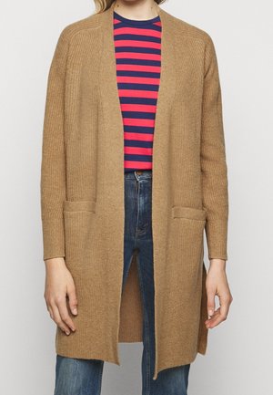 Nainen, jolla on päällään pitkä kamelinvärinen neuletakki punaisen ja laivastonsinisen raidallisen paidan sekä sinisten farkkujen päällä, seisoo yksiväristä taustaa vasten.