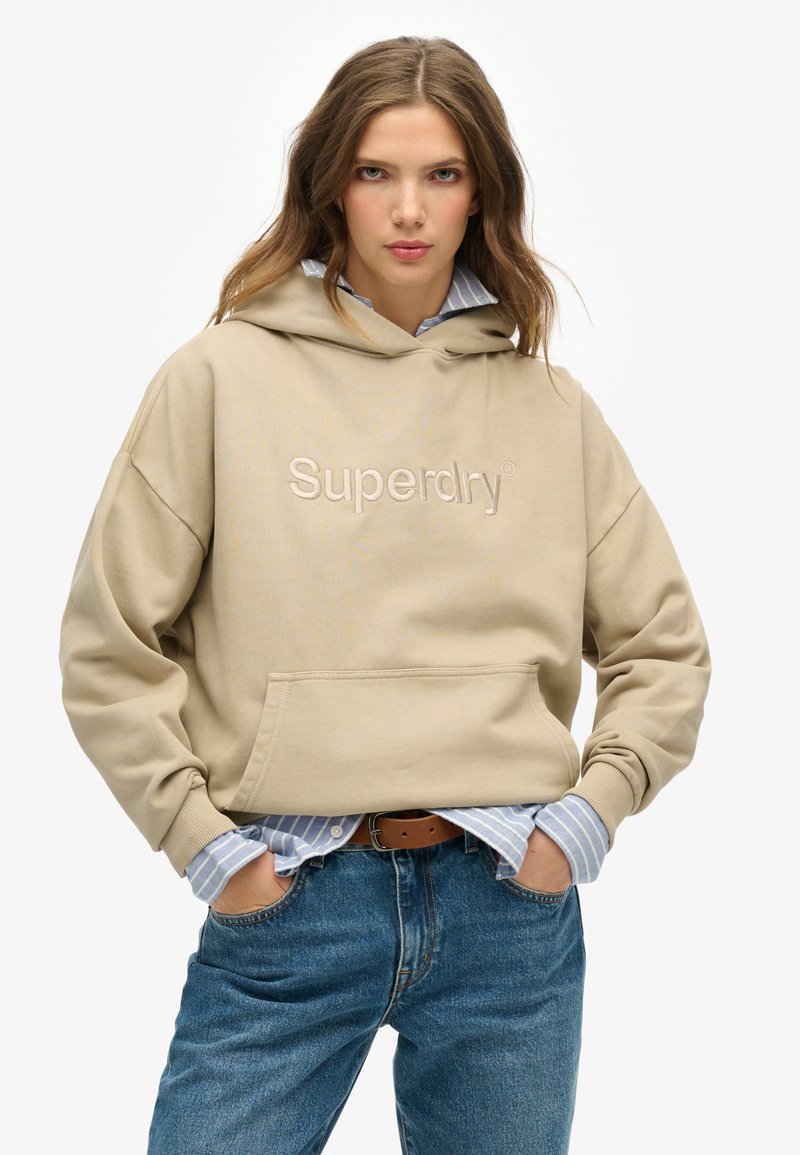 Beige hoodie met het logo "Superdry", voorzak en een relaxte pasvorm, gedragen over een blauw gestreept shirt en gecombineerd met spijkerbroeken.