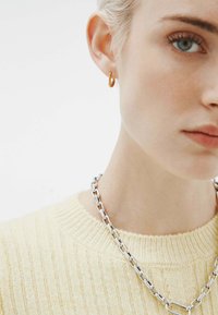 Aro de oro en la oreja de un modelo, combinado con un collar de cadena de plata. El modelo lleva un suéter amarillo de canalé con cuello redondo.