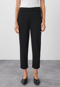 Černé, ušité kalhoty s hladkou texturou, délky po kotníky, elastickým pasem a jemnými postranními rozparky. Kombinováno s černými špičatými botami.