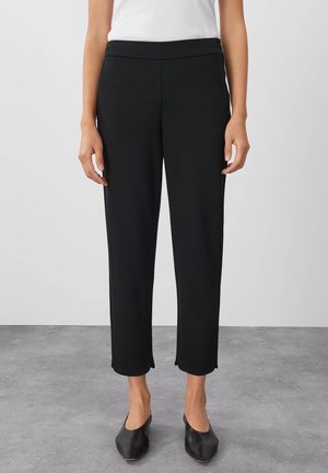 Pantalons noirs ajustés avec une texture lisse, longueur cheville, taille élastique et fentes latérales subtiles. Assortis à des chaussures noires à bouts pointus.