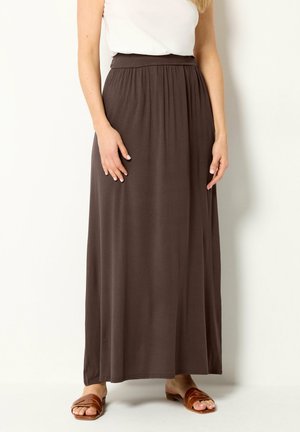 Kobieta w brązowej długiej spódnicy z wysokim stanem, białej bluzce bez rękawów i brązowych sandałach z odkrytymi palcami na tle jednolitej ściany.