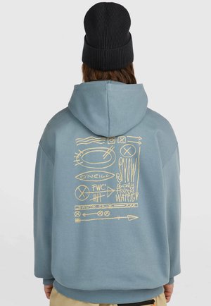 Person trägt einen hellblauen Hoodie mit gelber abstrakter Grafik und Text auf dem Rücken und eine schwarze Strickmütze.