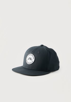Svart baseballcap med flat brem og en rund hvit logo-patch med teksten "J. Lindeberg Stockholm" på frontpanelet.