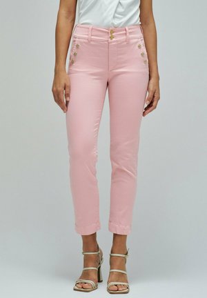 Mujer con pantalones cortos de talle alto en rosa claro con botones dorados en los laterales y cierre de doble botón, combinados con tacones beige de tiras.