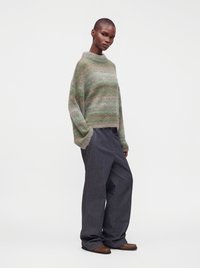 Personne portant un pull ample rayé vert et rose, un pantalon large gris foncé et des chaussures marron, debout devant un fond blanc uni.