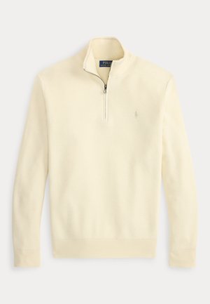 Homme portant un pull crème à fermeture éclair quart de zip, une chemise bleue à col, un jean foncé, une ceinture marron, des mocassins marron et une montre-bracelet, debout les mains dans les poches.