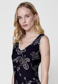 Robe sans manches bleu marine avec broderies florales en blanc et rouge, présentant un décolleté en V et un ourlet en forme de coquille le long du col.