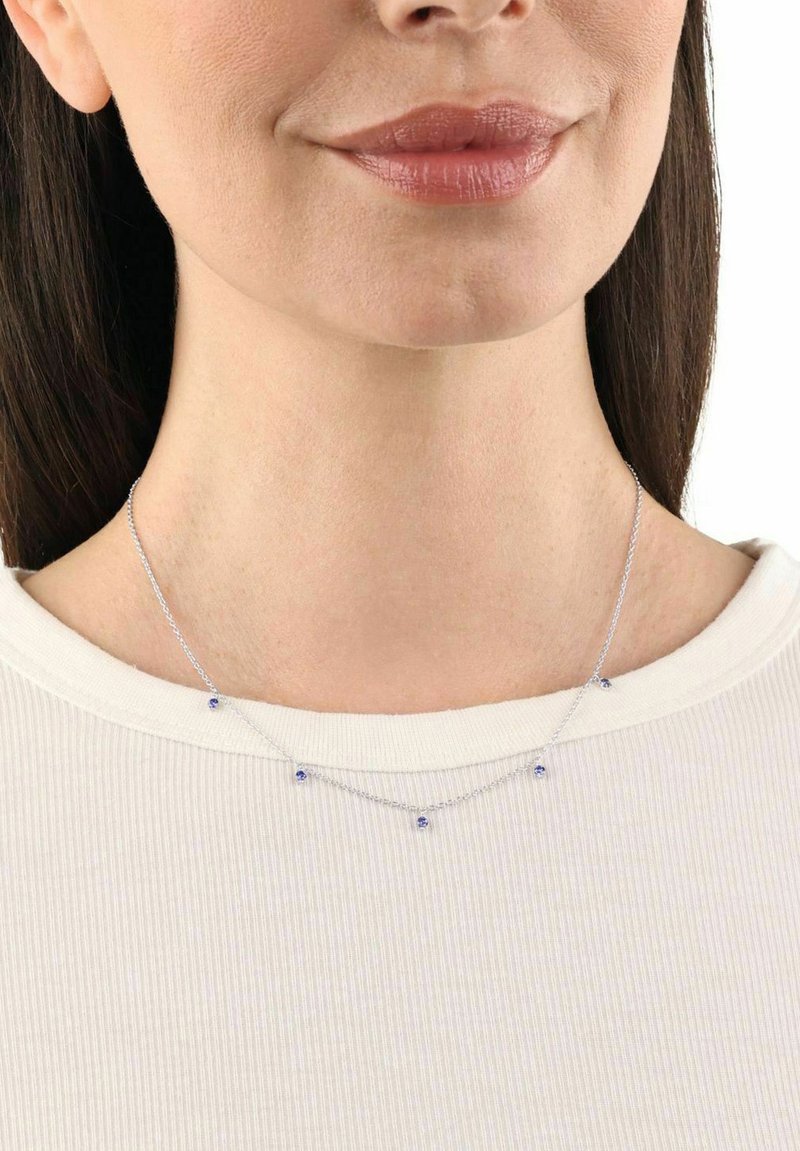Collar de plata que presenta pequeñas piedras preciosas azules, diseñado delicadamente con una cadena y múltiples acentos. Lleva puesto alrededor del cuello, sobre una blusa blanca.