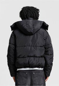 Sort pufferjakke med hætte, der har quiltet design, skinnende overflade og elastisk kant. Bagfra ses en afslappet pasform og minimal udsmykning.