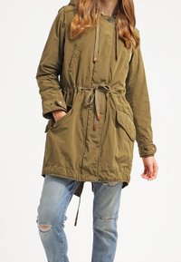 Mujer vistiendo un abrigo con capucha en verde oliva y cintura ajustable, con jeans azules claros desgastados, de pie contra un fondo blanco liso.