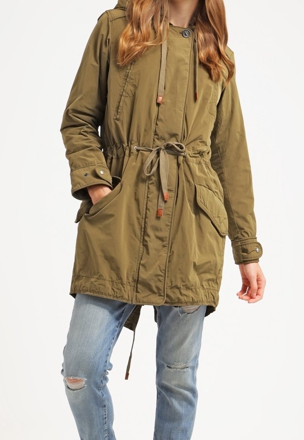 Mujer vistiendo un abrigo con capucha en verde oliva y cintura ajustable, con jeans azules claros desgastados, de pie contra un fondo blanco liso.