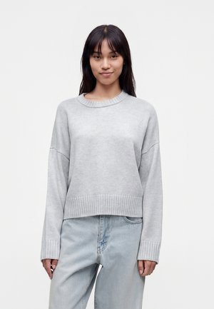 Jeune femme aux longs cheveux foncés portant un pull côtelé gris clair et un jean bleu clair ample sur un fond blanc uni.