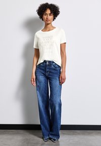 Hvid T-shirt med tekst i et centralt design, kombineret med brede blå denimjeans. Jeansene har en lys vask og to forlommer.