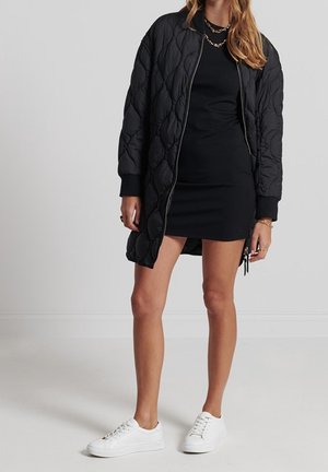 Femme portant une longue veste matelassée noire sur une robe noire avec des colliers dorés et des baskets blanches, debout devant un fond uni.