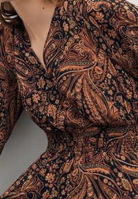 Robe noire avec des motifs en paisley orange et or délicats, taille ajustée, manches longues et décolleté en V. Texture de tissu douce.