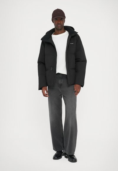 Black puffer jacket with a hood, featuring a front zipper and a patch pocket. Worn over a white shirt, paired with gray jeans and black shoes.