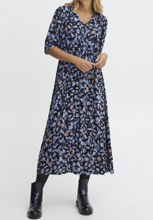 Femme portant une robe midi noire à fleurs bleues et roses, associée à des bottines noires, debout devant un fond blanc.