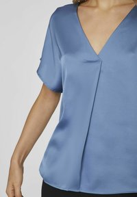 Blouse en satin bleu avec un décolleté en V, des manches courtes et une texture douce, présentant un léger pli décoratif pour un intérêt de design supplémentaire.