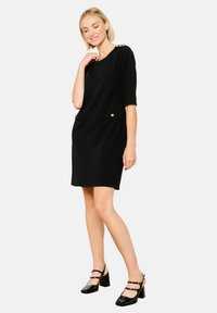 Vestido negro de manga corta con escote redondeado, que presenta acentos de botones dorados y bolsillos en los laterales. Hecho de una tela suave, hasta la rodilla.