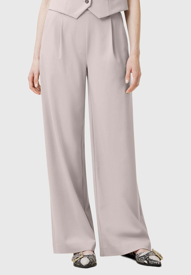 Pantaloni larghi grigi chiari realizzati in tessuto morbido con pieghe frontali, abbinati a scarpe bianche e nere con motivo e una fibbia decorativa.