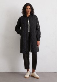Veste longue matelassée noire avec fermeture éclair à l'avant, poignets côtelés et poche latérale, associée à un pantalon noir cropped et des baskets beiges.