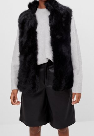 Personne portant une veste en fausse fourrure noire par-dessus un pull gris clair et un short en cuir noir jusqu'aux genoux, sur un fond uni.