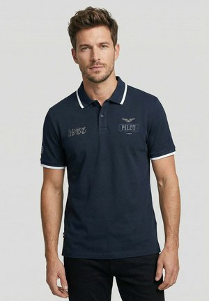 Homme portant un polo bleu marine avec bordure blanche, patchs brodés "1953" et "PILOTE" sur la poitrine, se tenant devant un fond uni.