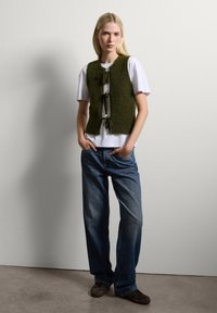 Olijfgroen gebreid mouwloos vest met een gestructureerde afwerking en voorste strikken, gedragen over een wit T-shirt, gecombineerd met blauwe wijde jeans.