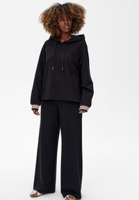 Czarna bluza z kapturem, z sznurkami, o luźnym kroju i szerokich rękawach, w zestawie z czarnymi spodniami o szerokich nogawkach. Uzupełniona o paskowane mankiety i wzorzyste buty.