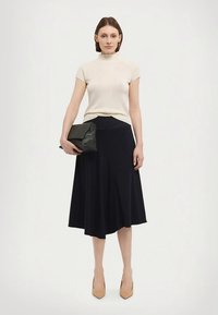 Béžový žebrovaný top s krátkými rukávy v kombinaci s tmavě modrou rozšířenou sukní. Modelka drží černé psaníčko. Outfit doplňují béžové lodičky.