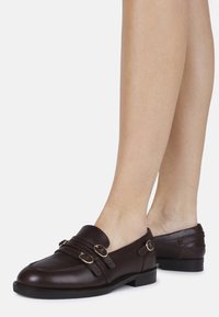 Dark brown leather loafers with two decorative straps and metal accents. Low heel and rounded toe. Smooth texture and minimalist design.