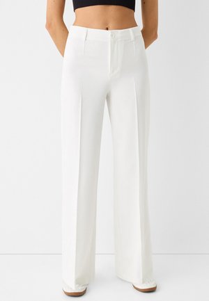Vrouw draagt hoge taille witte wijde broek met voren en een zwart cropped top, staand tegen een effen achtergrond.