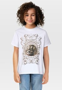 Biała bawełniana koszulka z nadrukiem w stylu vintage przedstawiającym dziewczynkę z delikatnymi lokami, otoczoną misternymi brązowymi wzorami i napisem: "Z powrotem do Hogwartu - Hermione."