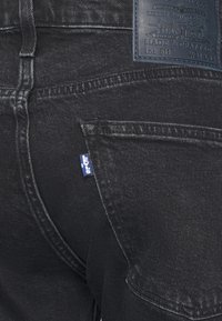 Jeans de mezclilla negra con un corte recto. Las características incluyen una pequeña etiqueta azul de Levi's en el bolsillo trasero y una etiqueta de cuero en la cinturilla.