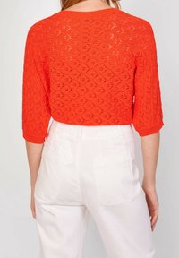 Punainen neulottu yläosa, jossa yksityiskohtainen kuvio, kolme neljäsosaa hihoissa ja pyöreä kaula-aukko, yhdistetty valkoisiin korkeavyötäröisiin housuihin.