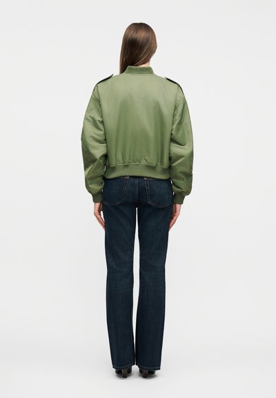 Άτομο με μακριά καστανά μαλλιά, που στέκεται με την πλάτη στραμμένη φορώντας μια φαρδιά μπουφάν bomber σε ελιά πράσινο και σκούρα μπλε τζιν με ίσια γραμμή.