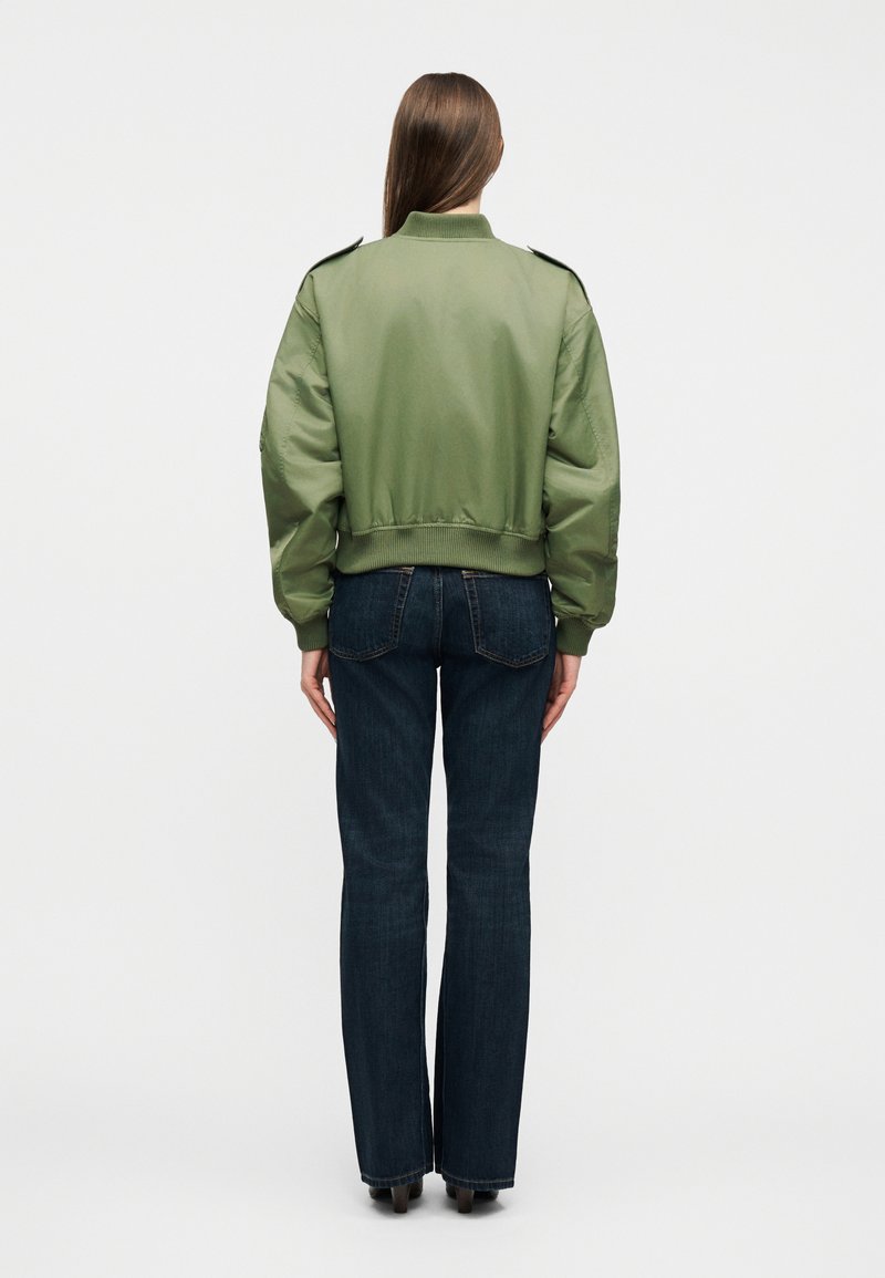 Personne aux longs cheveux bruns vue de dos, portant une veste bomber oversize vert olive et un jean droit bleu foncé.